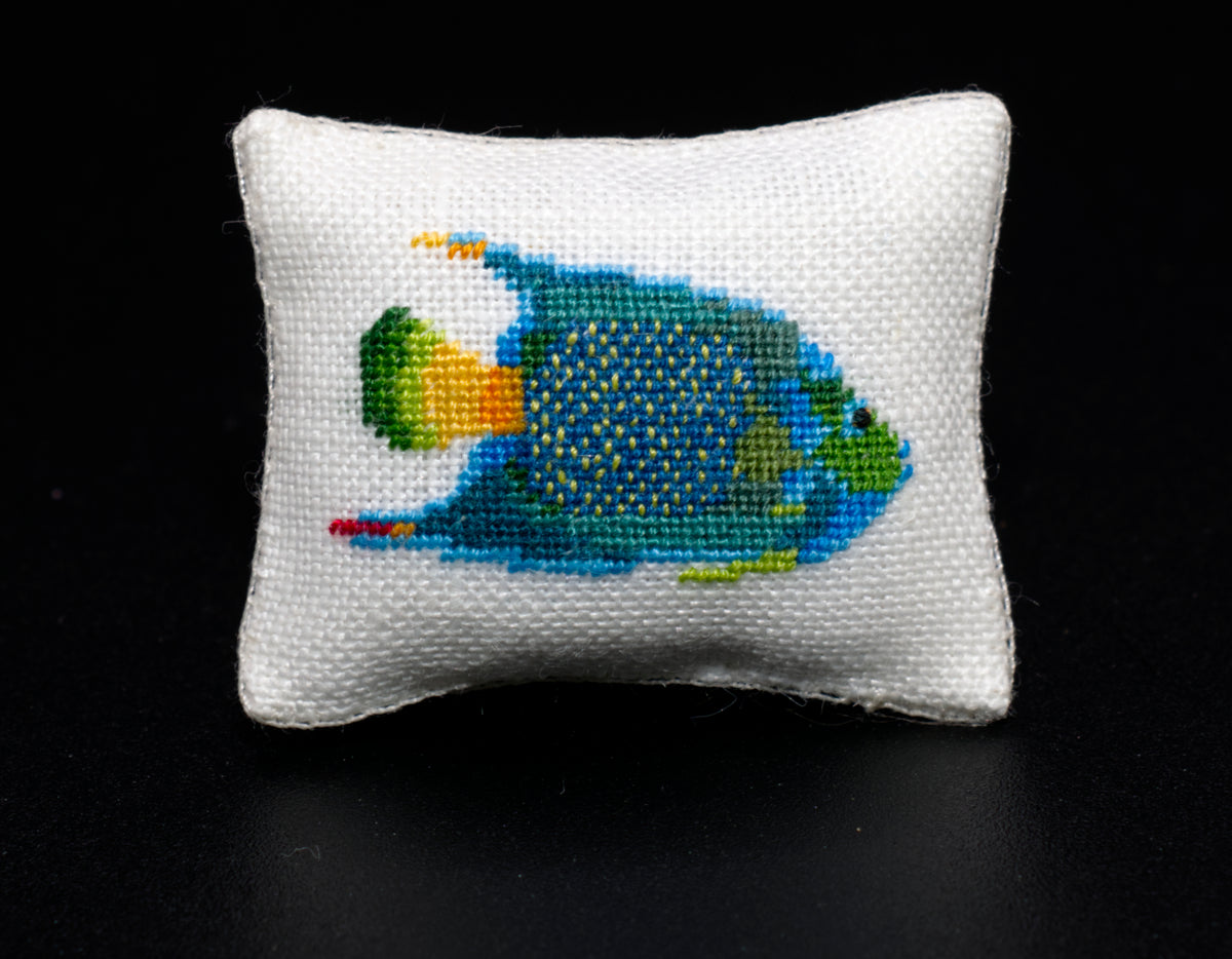 A small white petit point pillow featuring a tropical fish design in shades of blue, green, yellow, and red, set against a solid black background.