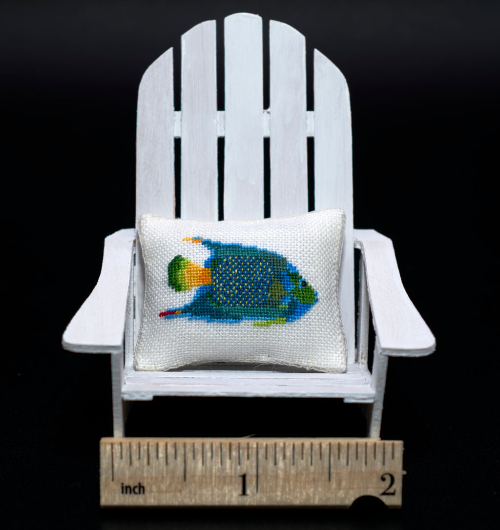 A miniature white Adirondack chair holding a small petit point pillow with a colorful fish design, with a wooden ruler at the bottom for scale against a black background.