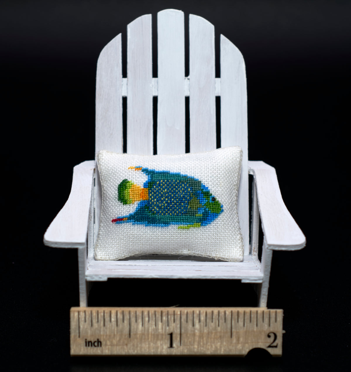 A miniature white Adirondack chair holding a small petit point pillow with a colorful fish design, with a wooden ruler at the bottom for scale against a black background.