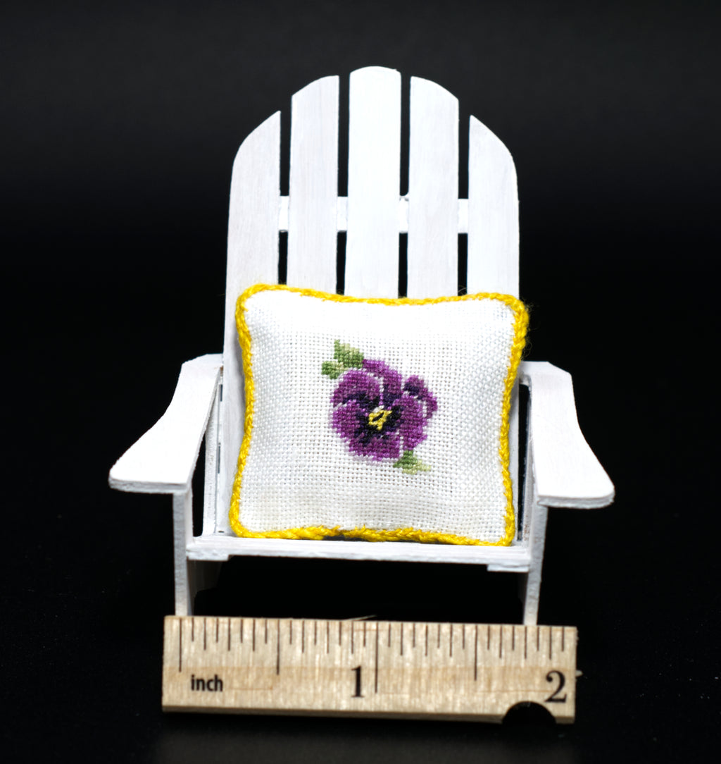 A miniature white Adirondack chair holding a small petit point pillow with a purple pansy and yellow border, shown with a wooden inch ruler at the bottom for scale against a black background.