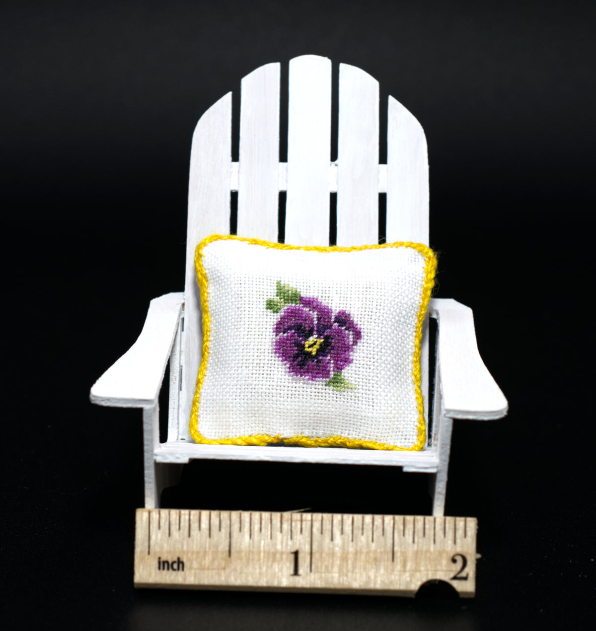 A miniature white Adirondack chair holding a small petit point pillow with a purple pansy and yellow border, shown with a wooden inch ruler at the bottom for scale against a black background.