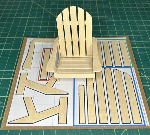 A miniature wooden Adirondack chair made from light-colored wood, displayed on top of a printed cardboard template sheet with matching cutout pieces on a green cutting mat.