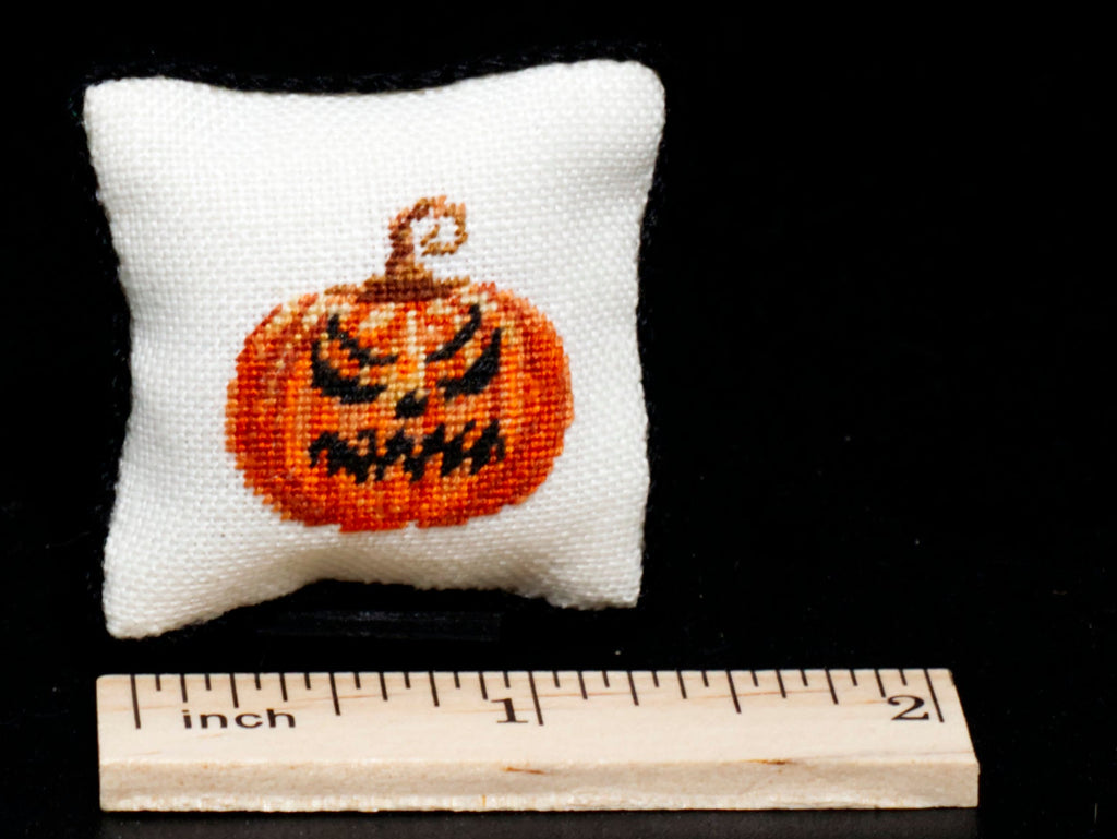 A small white square pillow with an evil-faced orange jack-o'-lantern in petit point on the front, shown next to a wooden inch ruler against a black background.