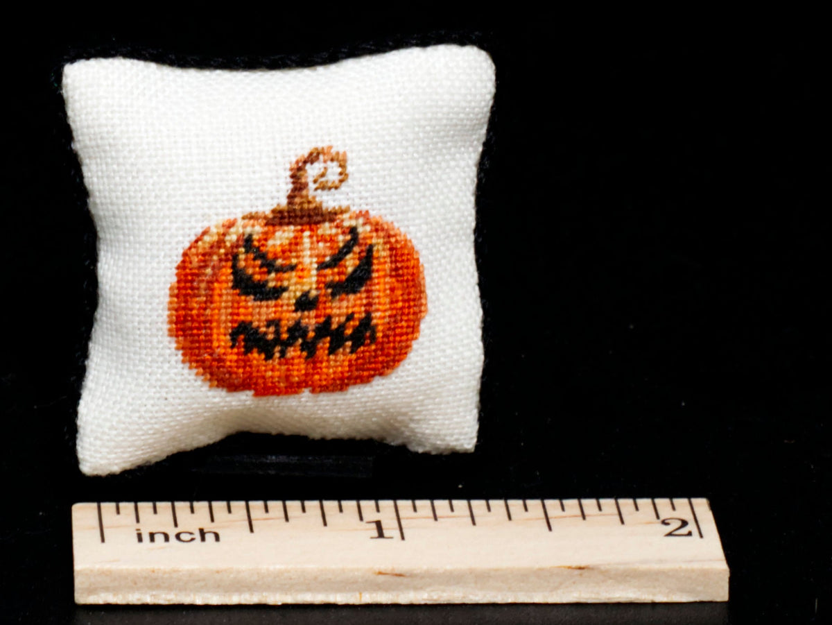 A small white square pillow with an evil-faced orange jack-o'-lantern in petit point on the front, shown next to a wooden inch ruler against a black background.