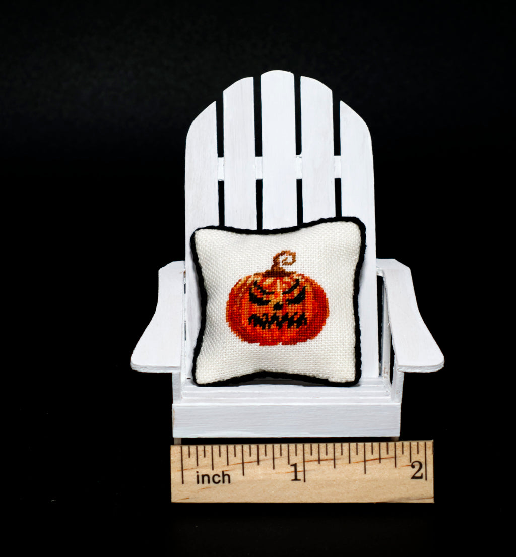 A small white petit point pillow with an evil orange jack-o'-lantern on the front, shown next to a wooden inch ruler against a black background.