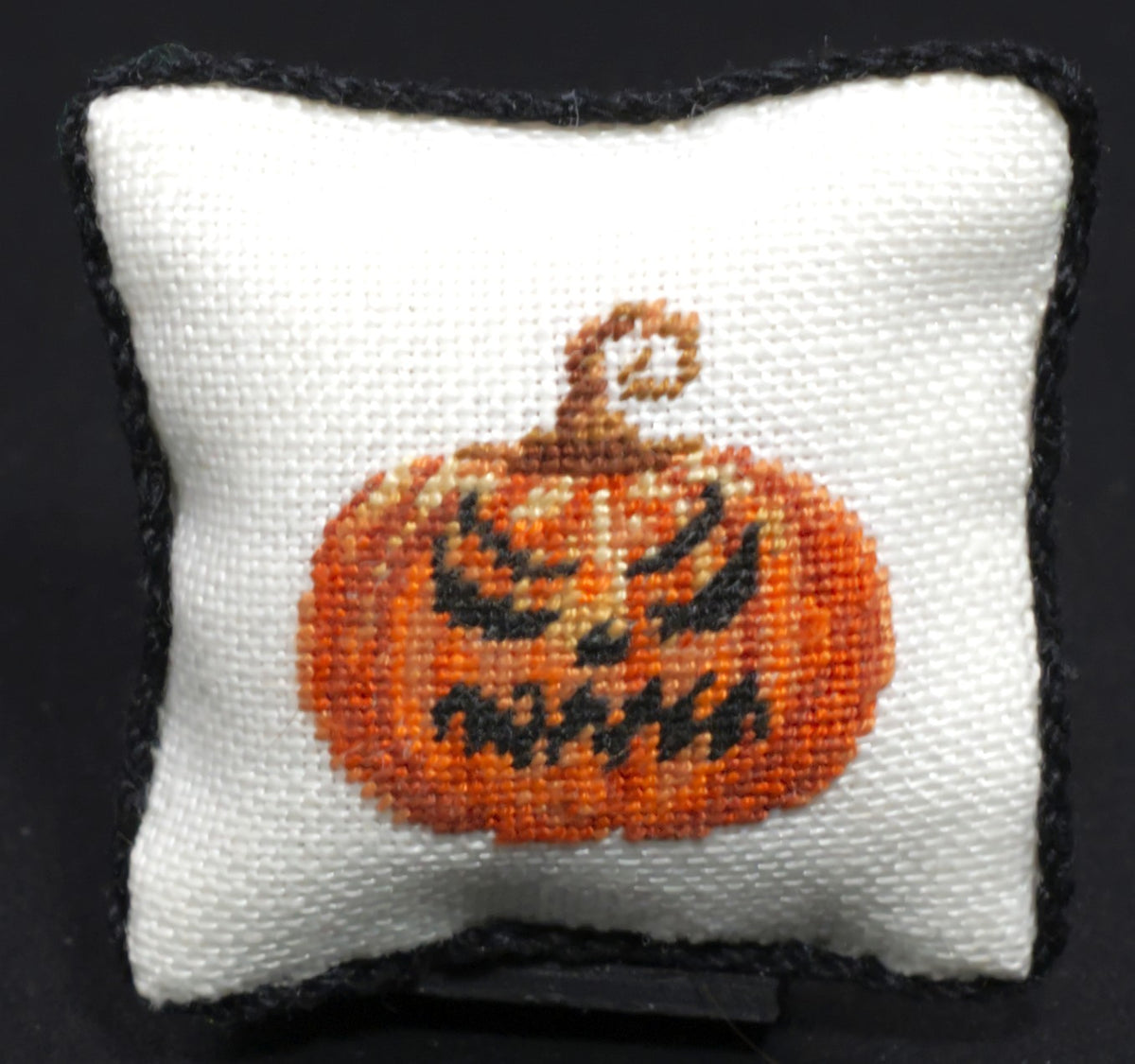 A square white petit point pillow with a black border and an evil orange jack-o'-lantern pumpkin design with a stitched stem, shown against a black background.