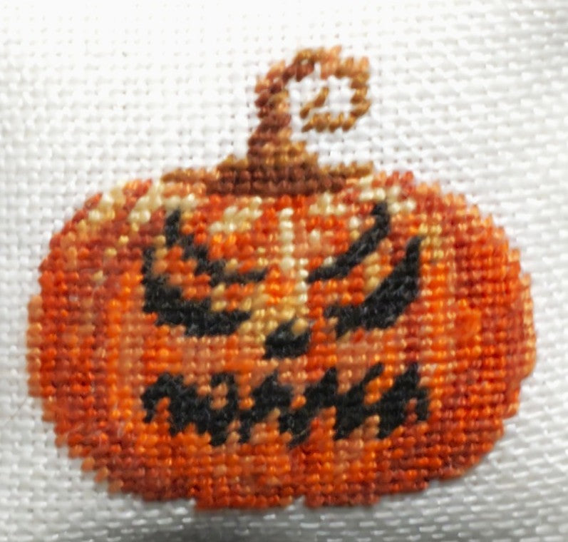 A close-up of a small white petit point pillow with an evil orange jack-o'-lantern on the front.