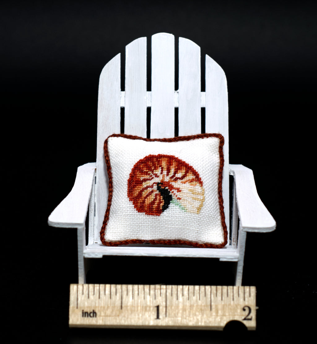 A miniature white Adirondack chair holding a small petit point pillow with a nautilus shell design, shown with a wooden ruler at the bottom for scale against a black background.