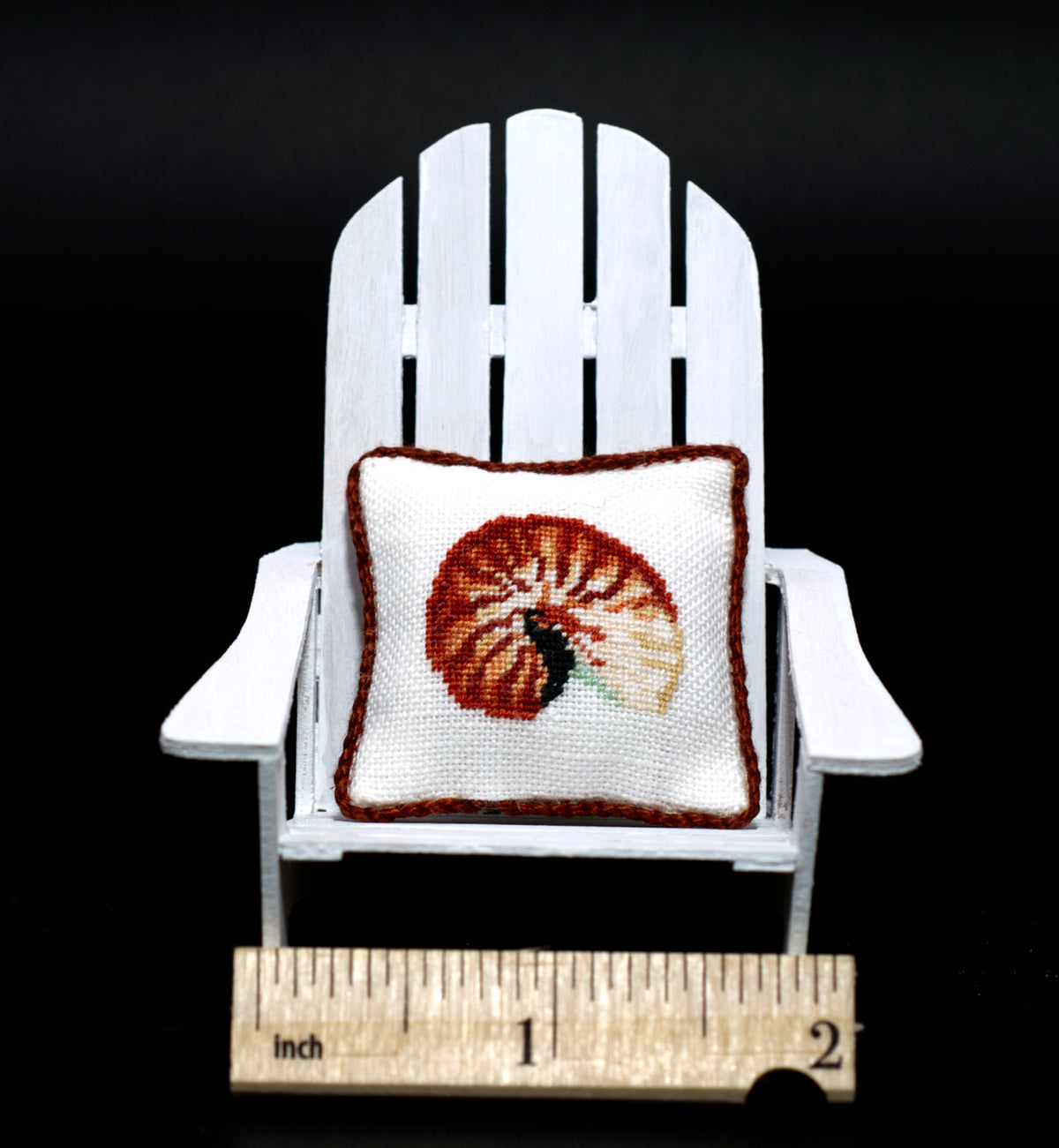 A miniature white Adirondack chair holding a small petit point pillow with a nautilus shell design, shown with a wooden ruler at the bottom for scale against a black background.