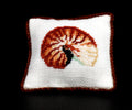 A square white petit point pillow with a brown border and a centered shell motif in orange, brown, black, and cream, shown against a solid black background.