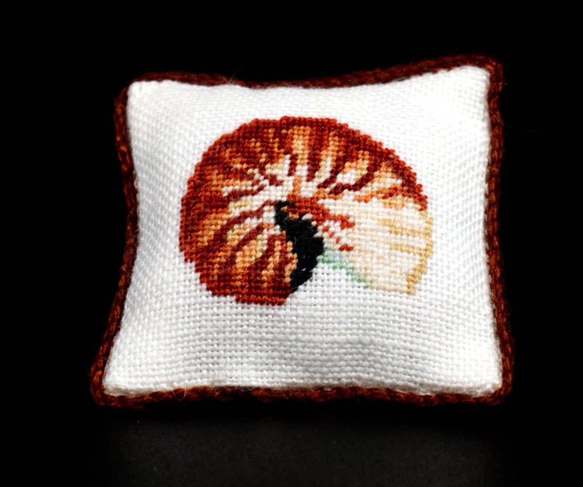 A square white petit point pillow with a brown border and a centered shell motif in orange, brown, black, and cream, shown against a solid black background.