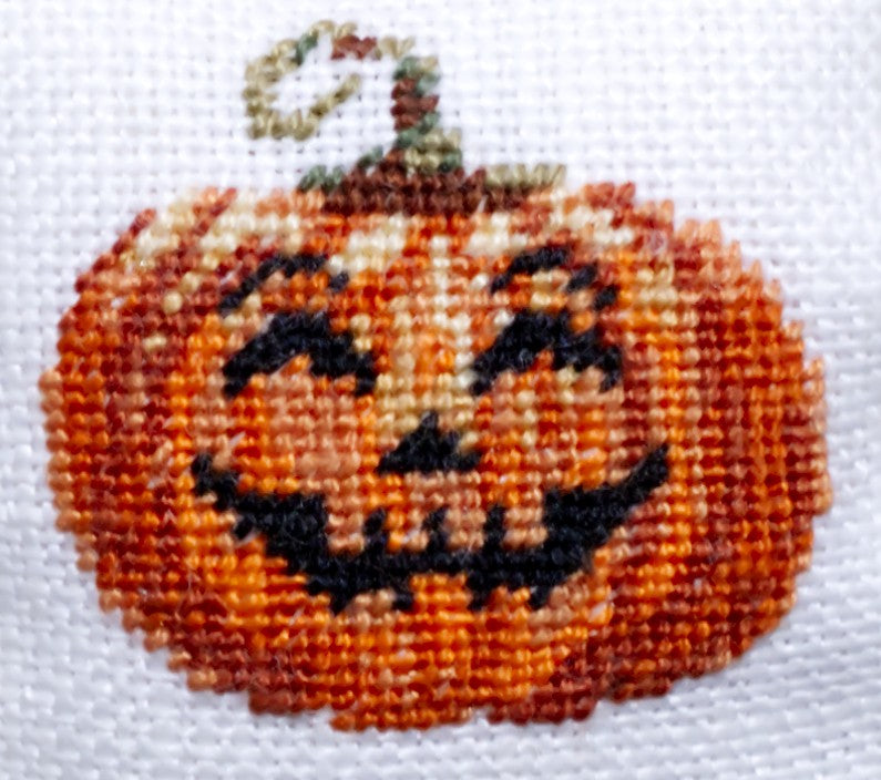 A close-up of a finished petit point design depicting a smiling jack-o'-lantern pumpkin on a white linen background