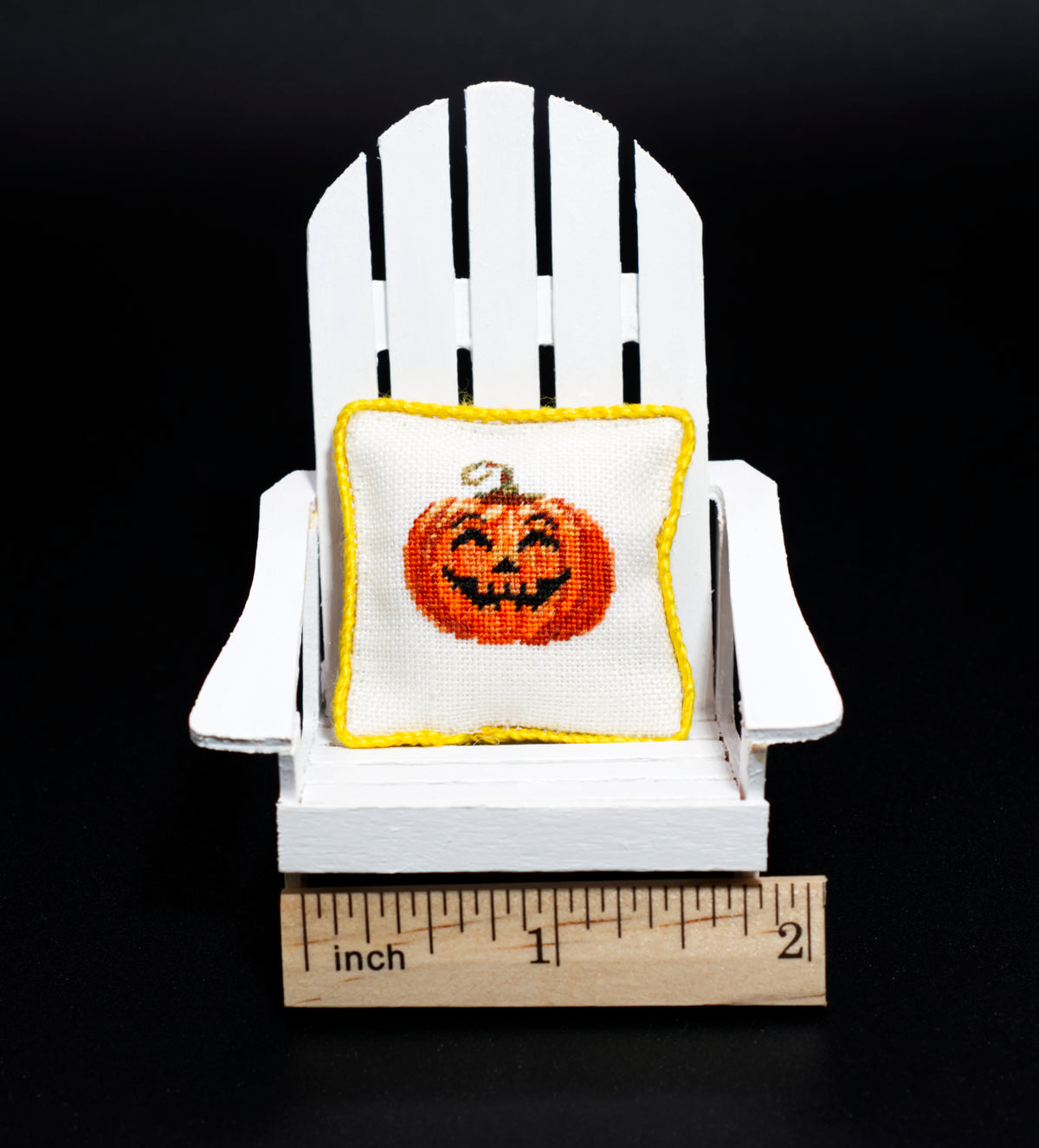 A miniature white Adirondack chair holding a small petit point pillow with a smiling jack-o'-lantern design, shown with a wooden ruler at the base for scale against a black background