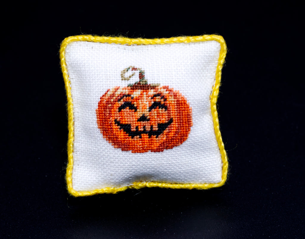 A small square white petit point pillow with a yellow border, featuring a smiling orange jack-o'-lantern face on the front, set against a solid black background.