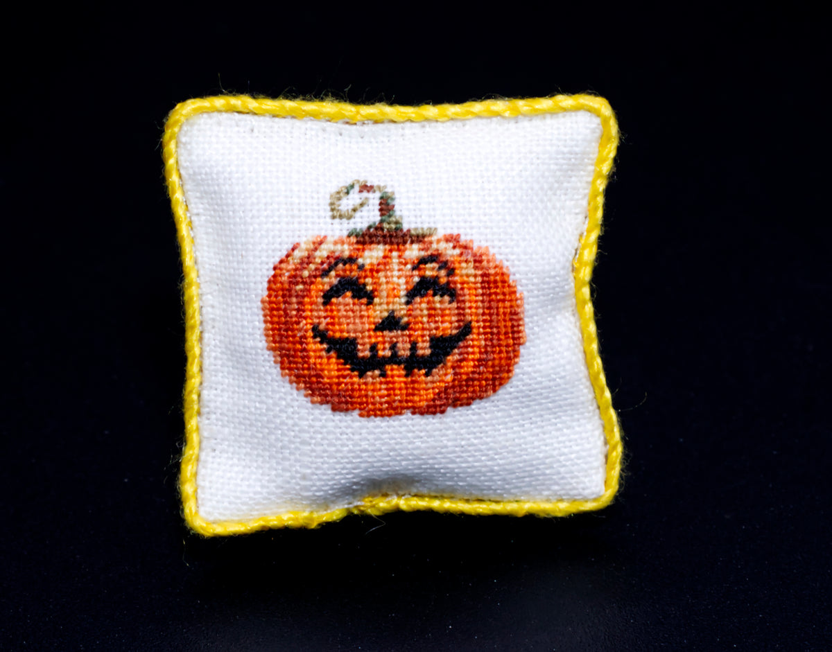 A small square white petit point pillow with a yellow border, featuring a smiling orange jack-o'-lantern face on the front, set against a solid black background.