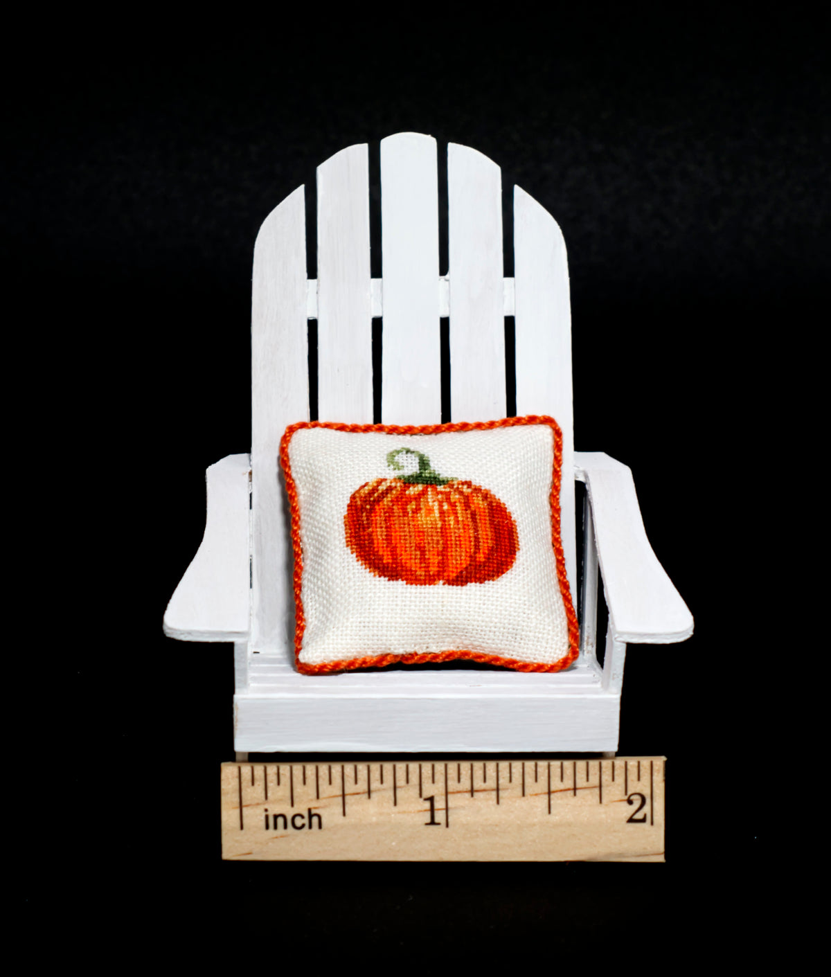 A small white Adirondack chair displayed against a black background, holding a miniature white pillow with an orange pumpkin design and orange border. A wooden inch ruler is placed in front of the chair to show scale.
