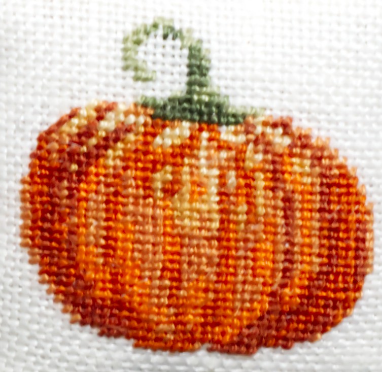 A close-up of a petit point pumpkin design on white linen fabric, featuring orange and copper stitched details with a green stem.