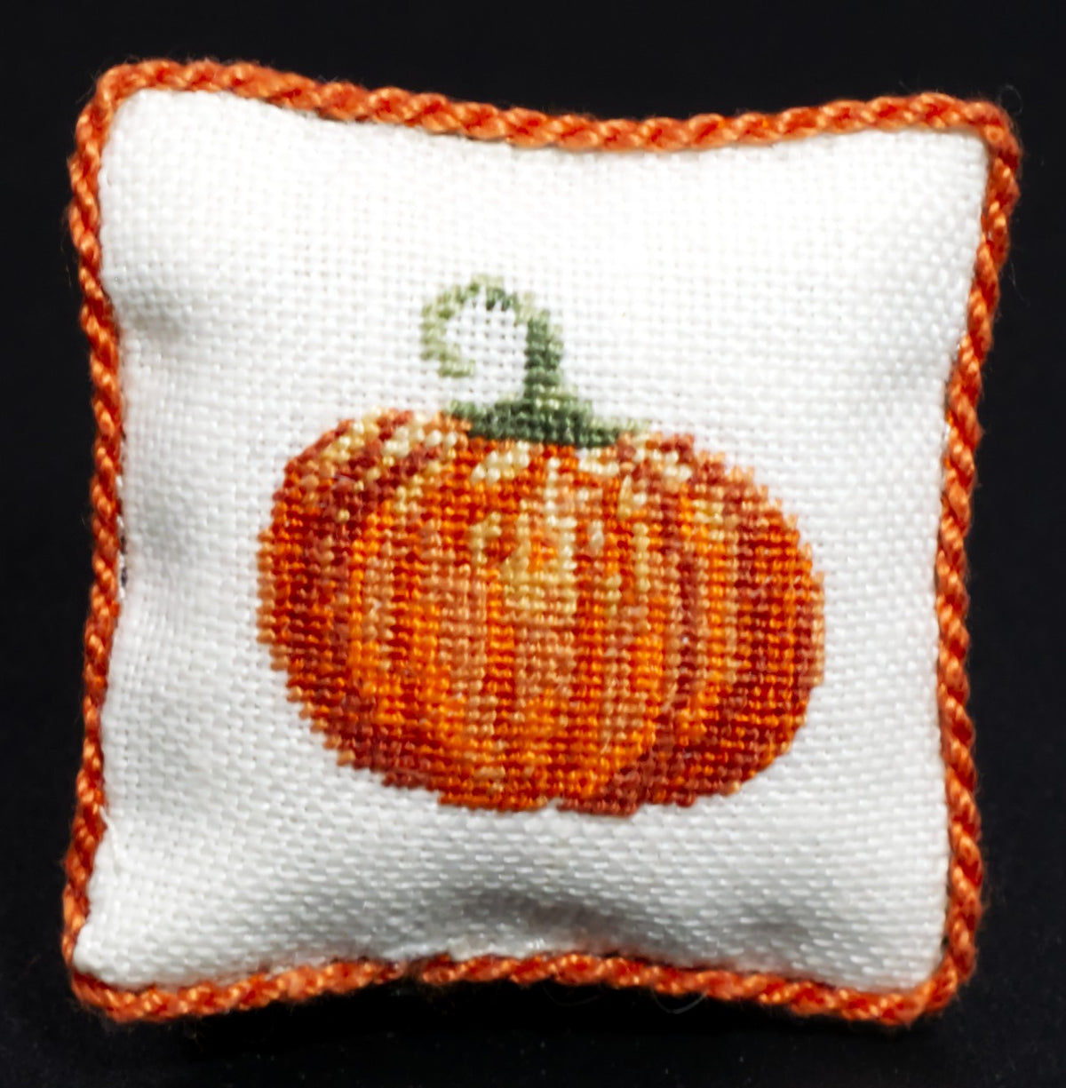 A square white petit point pillow with a braided orange border and a central orange pumpkin motif with a green stem, shown against a black background