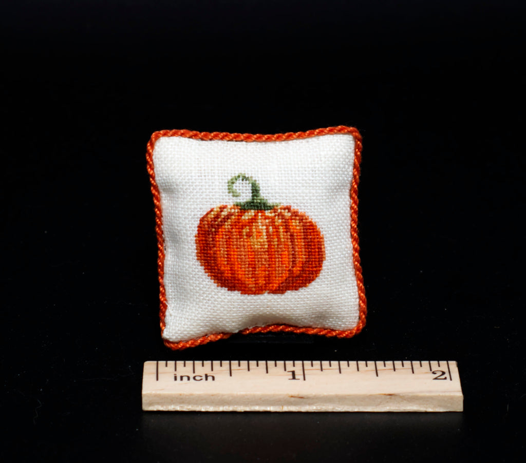 A small square white petit point pillow with an orange pumpkin design and orange braided border, shown on a black background with a wooden inch ruler below for scale.