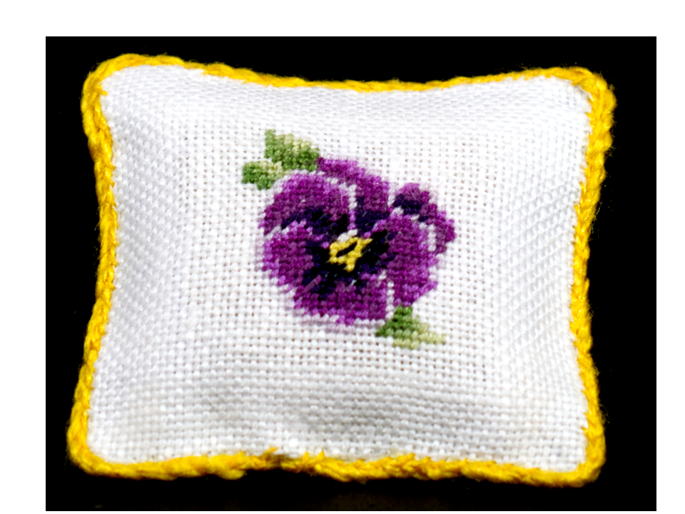 A square white petit point pillow with a bright yellow border and a central embroidered purple pansy flower with green leaves, set against a solid black background
