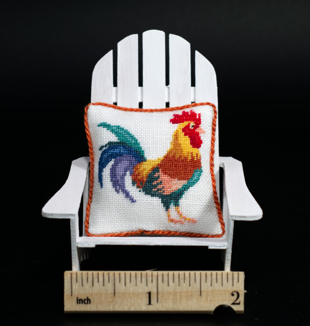 A small square petit point pillow with a colorful rooster design and orange border, displayed on a miniature white Adirondack chair against a black background, with an inch ruler below for scale.