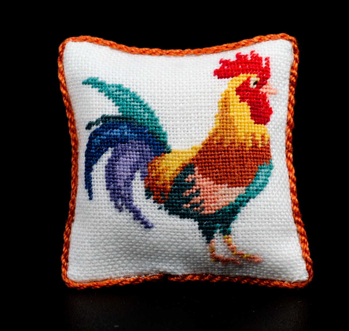 A square white petit point cushion with a colorful rooster design and an orange braided border, photographed against a solid black background.