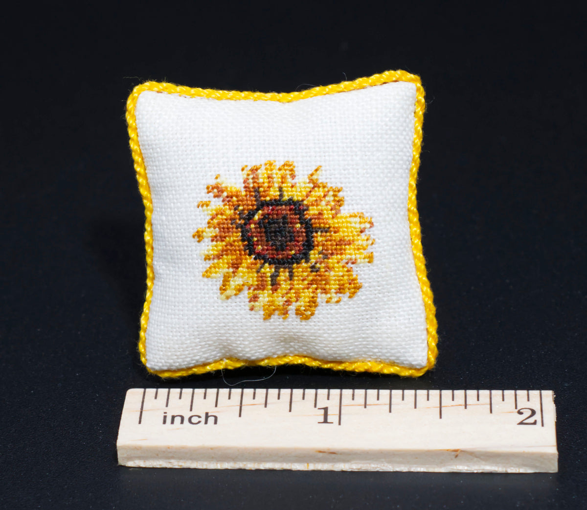 A square white fabric pillow with a yellow border and a petit point sunflower design in the center, shown above a wooden inch ruler on a black background.
