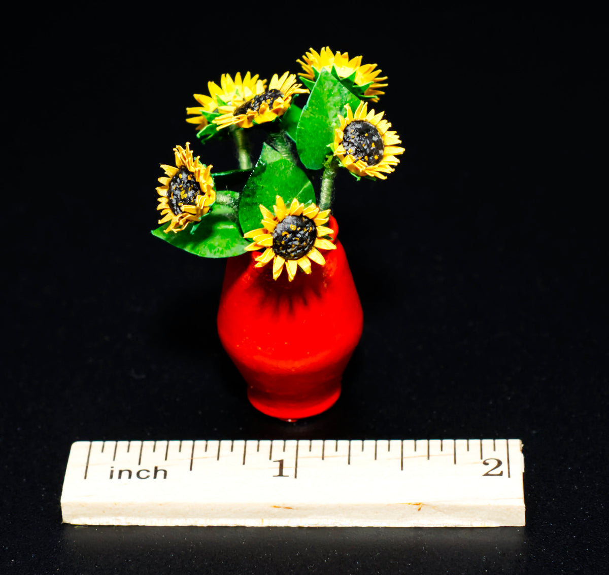 A miniature terra cotta vase holding a  bouquet of yellow sunflowers with green leaves with a wooden ruler at the bottom for scale.