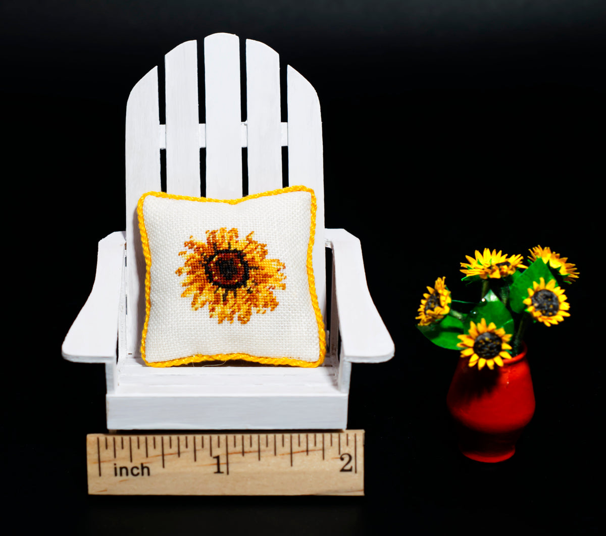 A miniature white Adirondack chair holding a small sunflower pillow, shown next to a vase of sunflowers and a wooden ruler for scale against a black background.