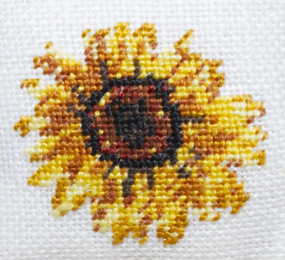 Close-up of a sunflower petit point design on antique white linen, featuring yellow, orange, brown, and dark brown petals with a dark center.